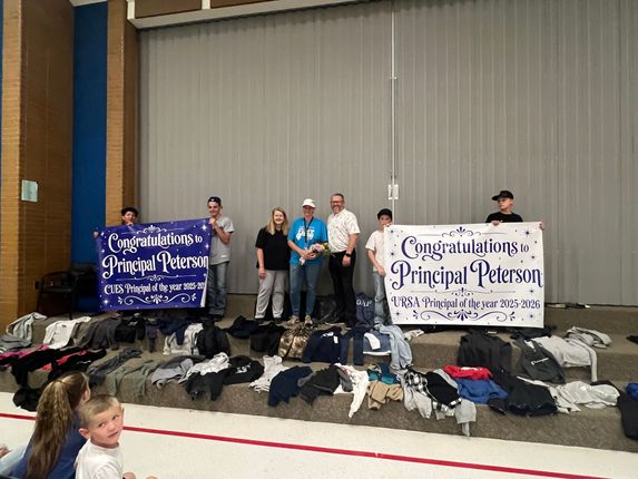 Group on stage with banners and piles of clothing, celebrating a principal program in a gymnasium.