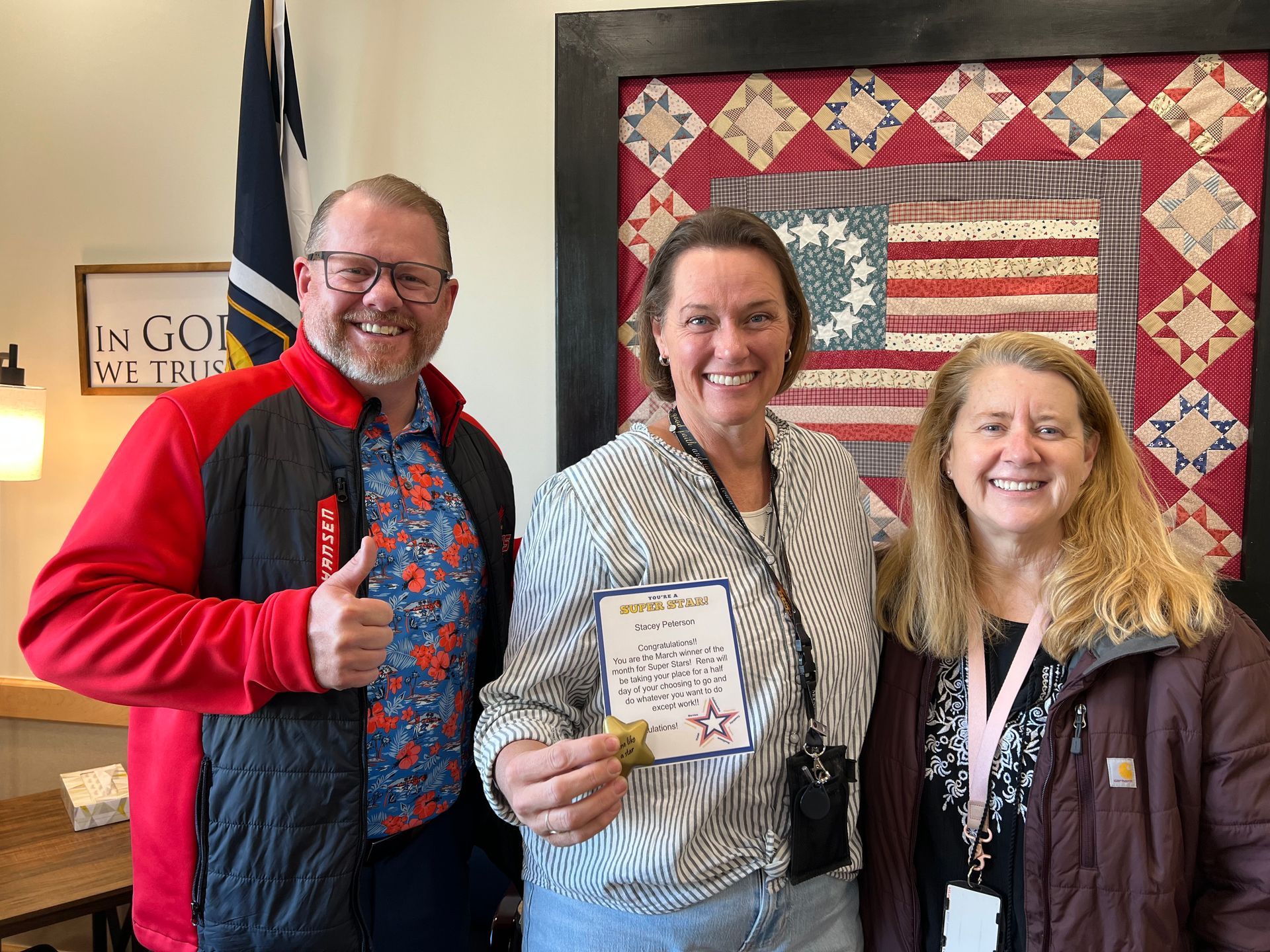 Three people smiling indoors, one holding an award certificate; quilt and flag in background