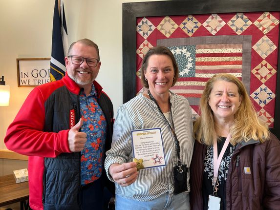 Three people smiling indoors, one holding a certificate, in front of a quilt display