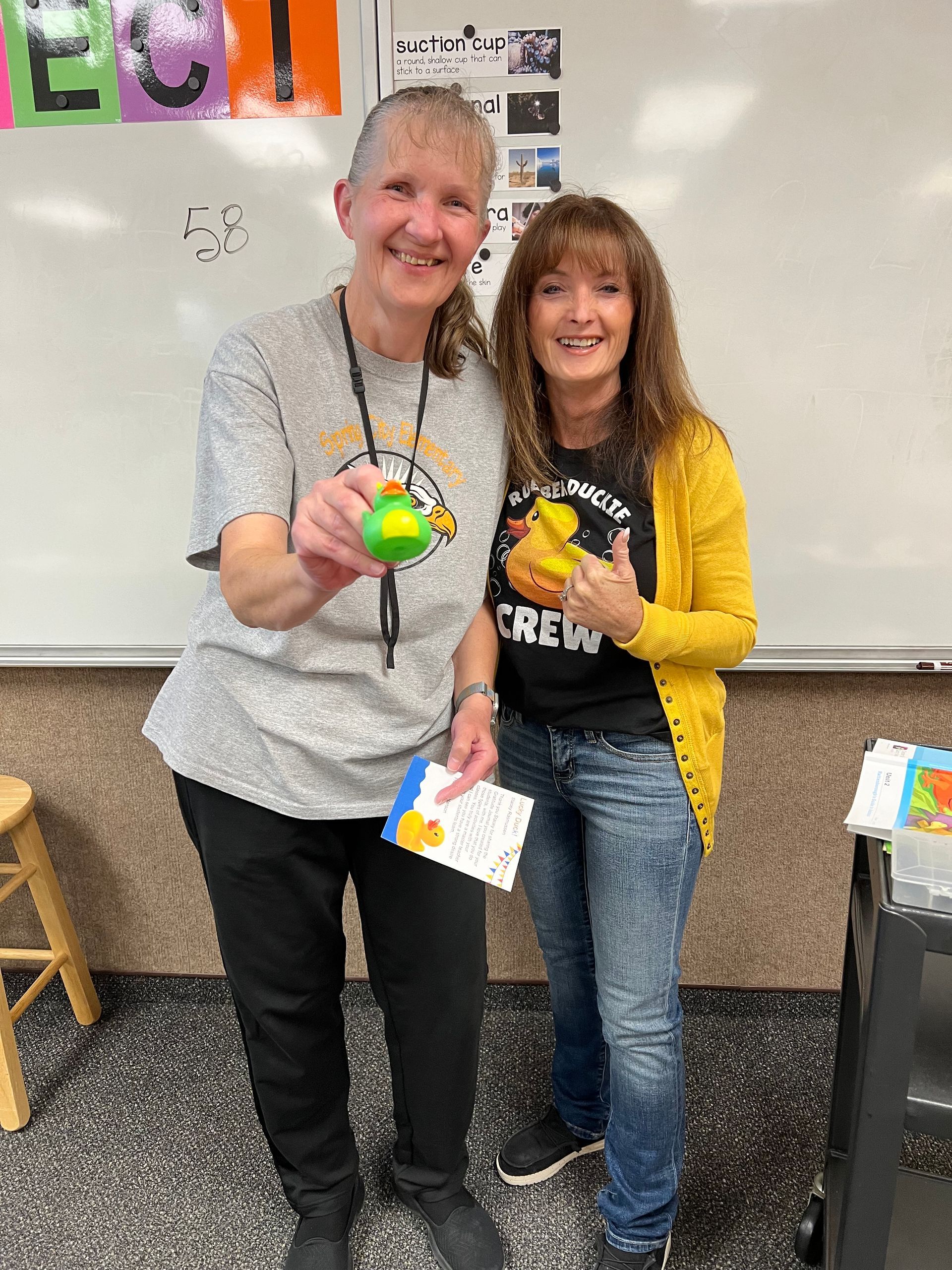 Two women are standing next to each other in a classroom holding rubber ducks.