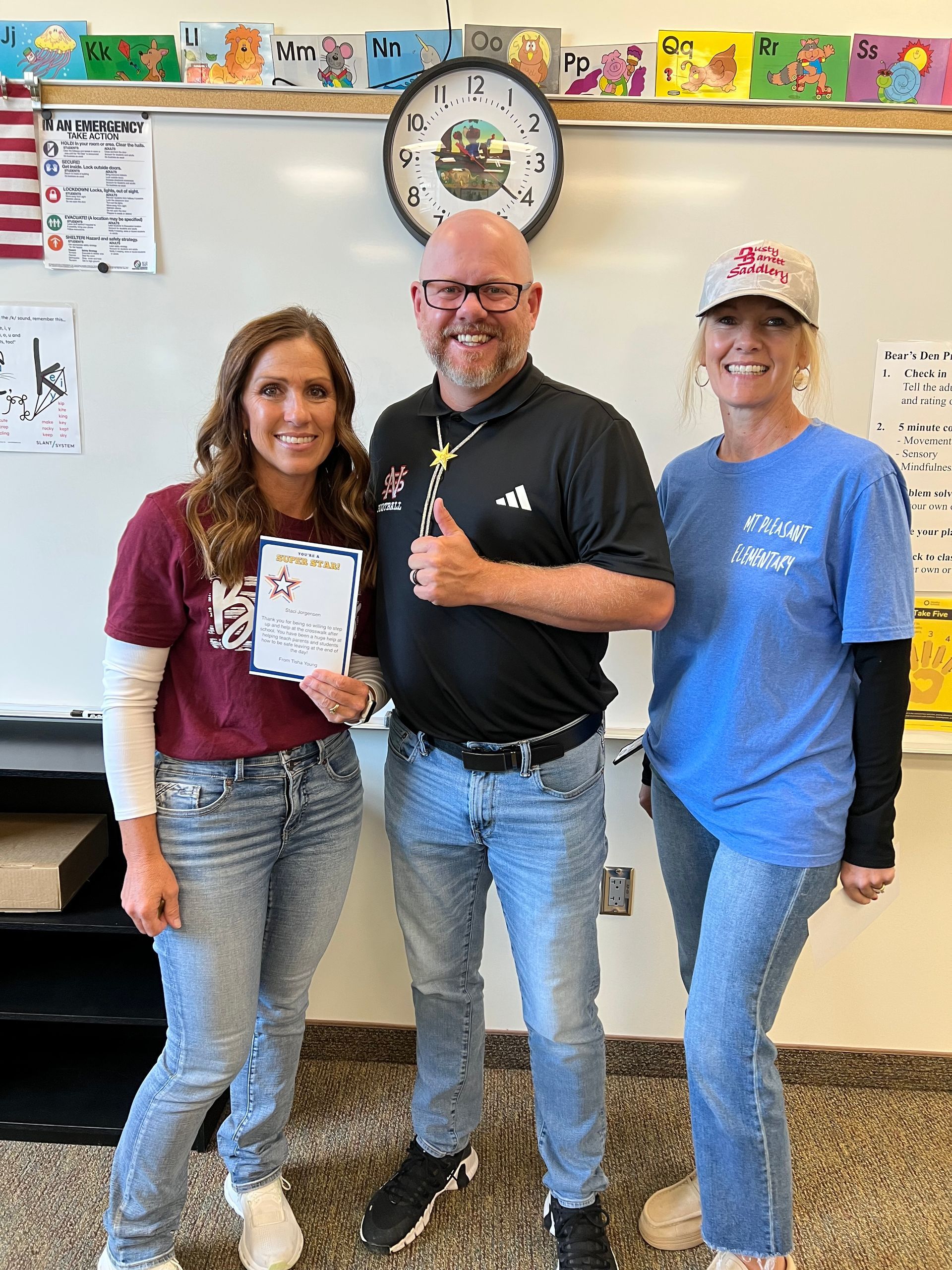 Three people standing in a classroom. Woman in maroon shirt holds paper. Man gives thumbs up, other woman smiles.