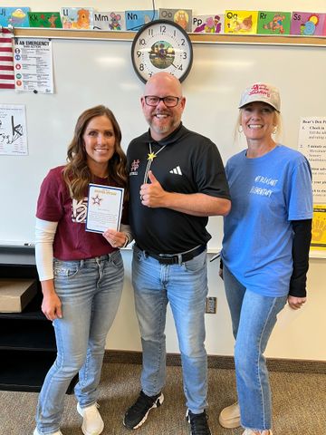 Three smiling people in a classroom, one holds an award. Man gives thumbs up.