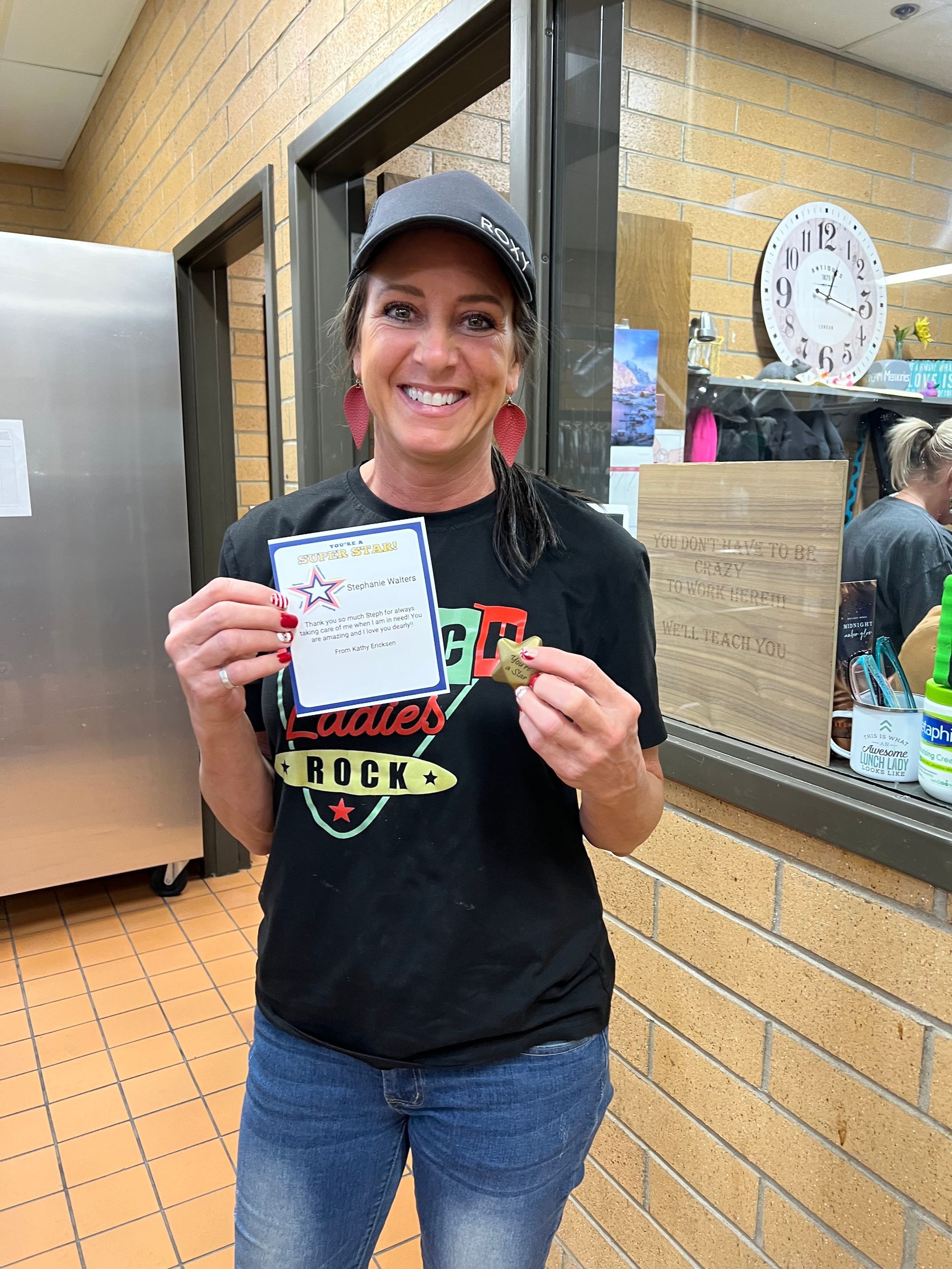 Woman in a cap and shirt, holding a note and a small item, smiling in a kitchen setting.