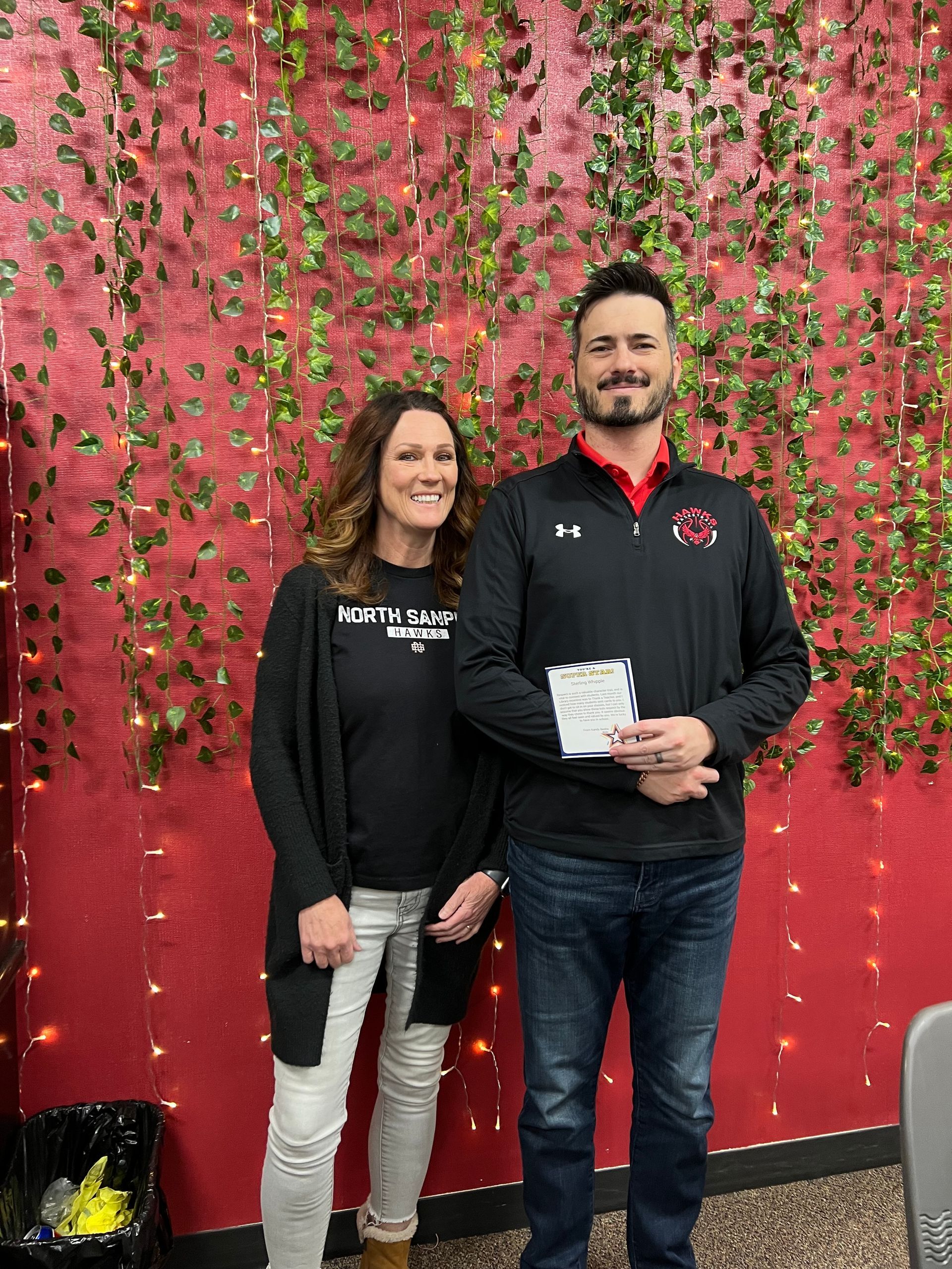 Woman and man stand together in front of a red wall decorated with greenery. The man holds a paper.