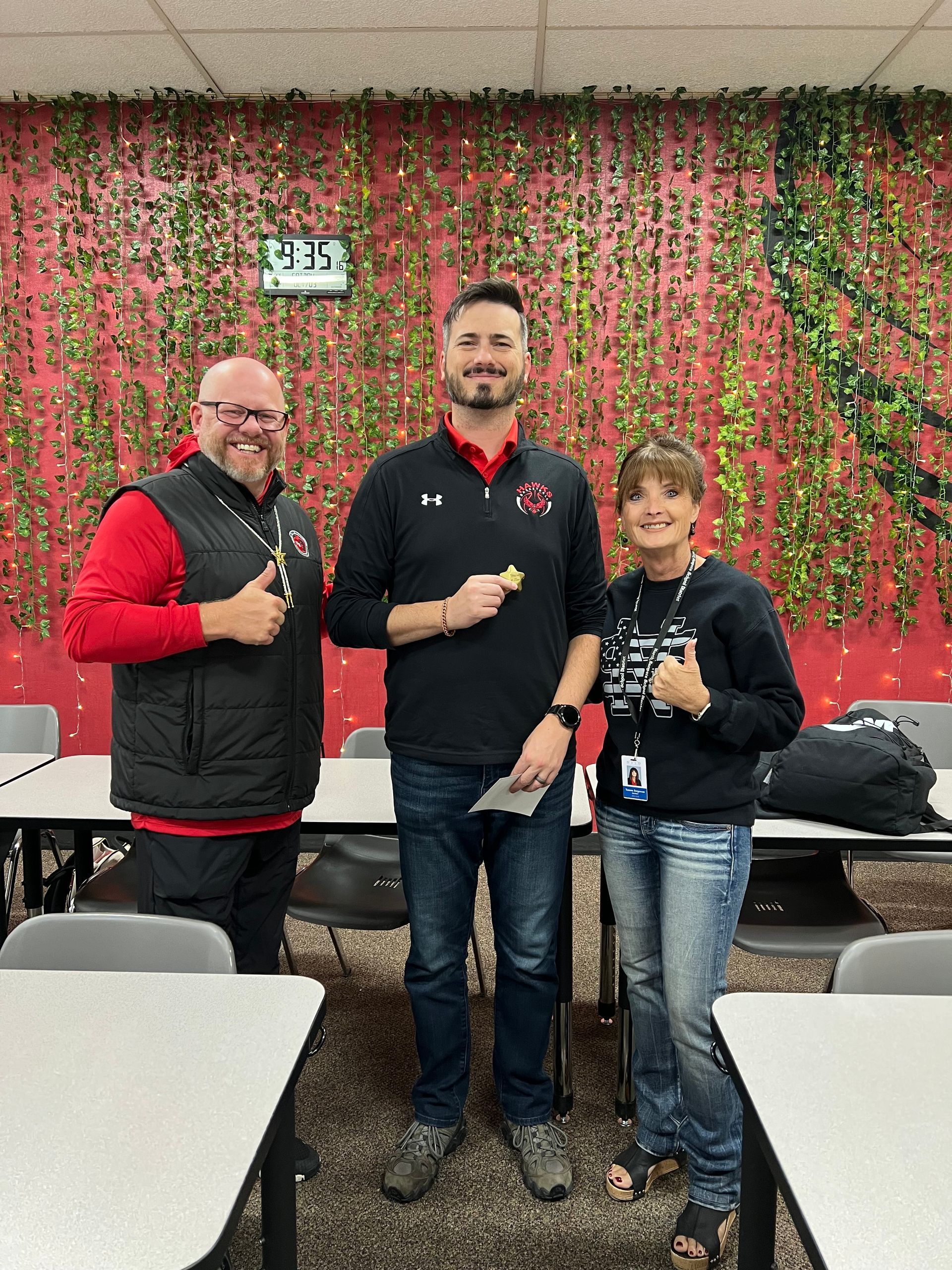 Three people, smiling and holding objects, stand in a classroom with a wall of ivy.