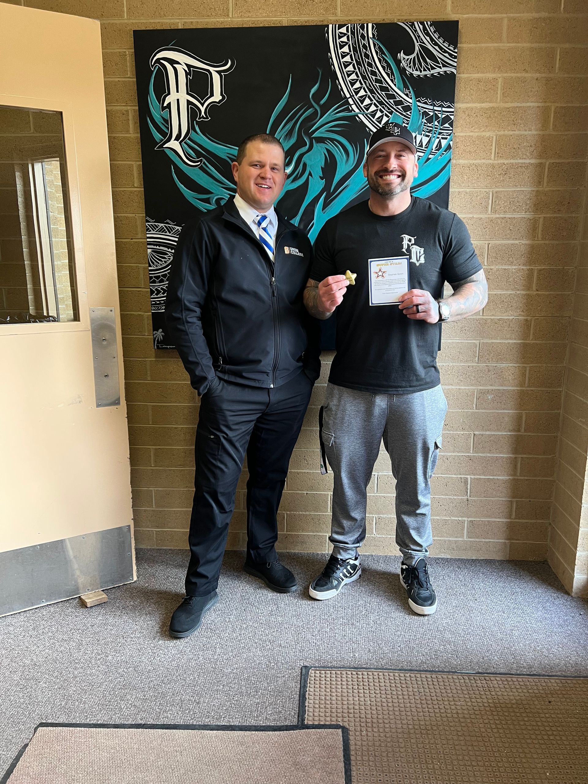 Two men stand indoors by a wall with artwork. One man holds a paper and a small object; both men smile.