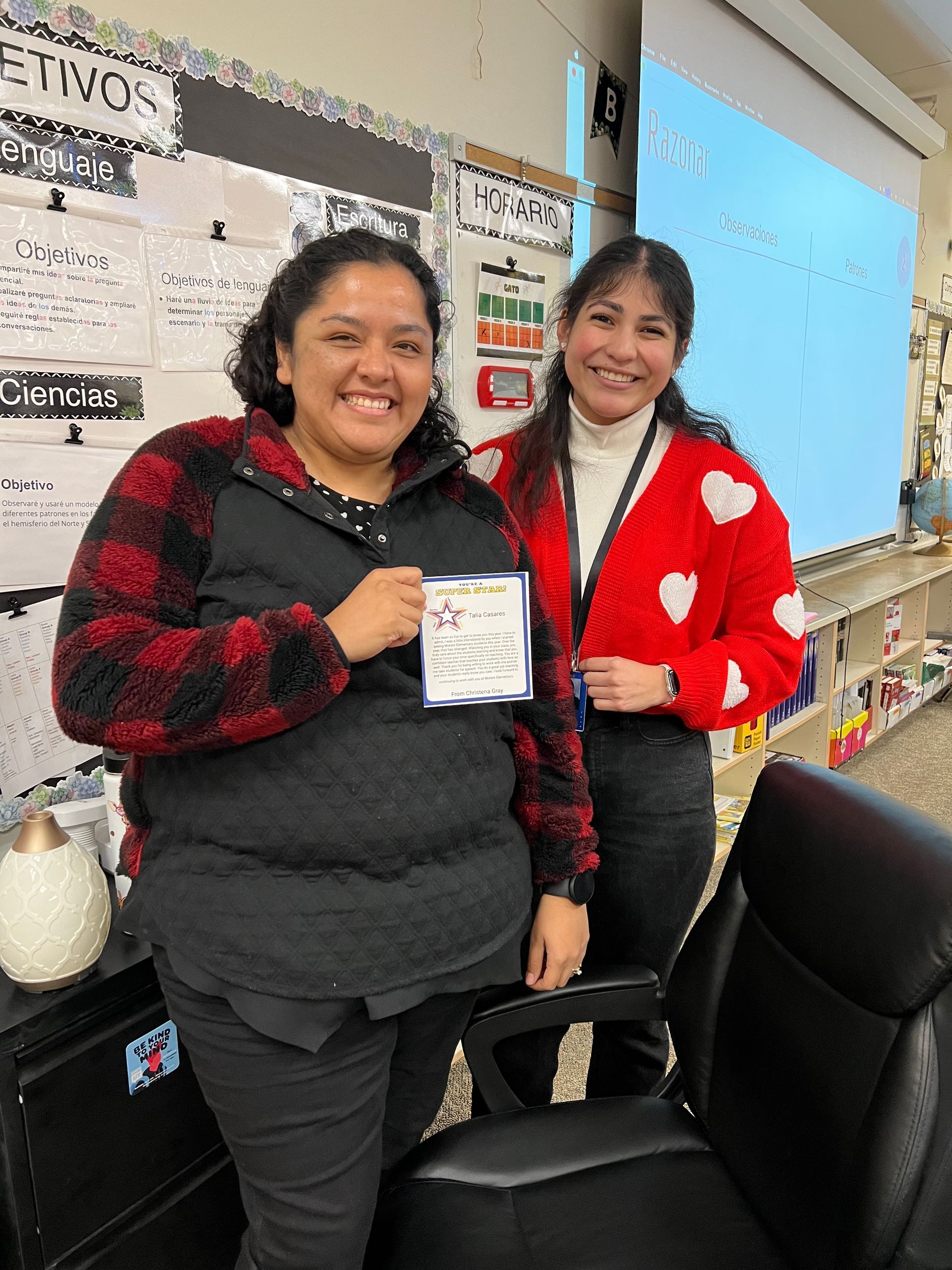 Two women smiling in a classroom, one holding a certificate.  One wears a red and black jacket, the other a red sweater with white hearts.