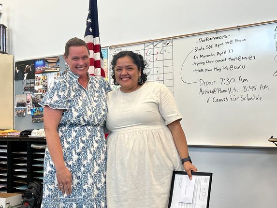 Two people smiling in a classroom, standing beside a whiteboard and holding a framed certificate.