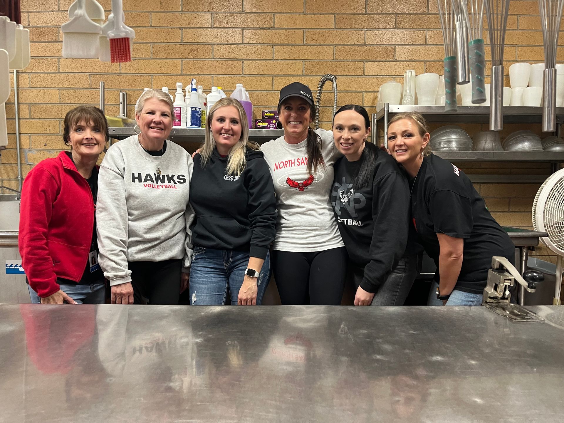 Six women smiling in a commercial kitchen with stainless steel surfaces and cleaning supplies.