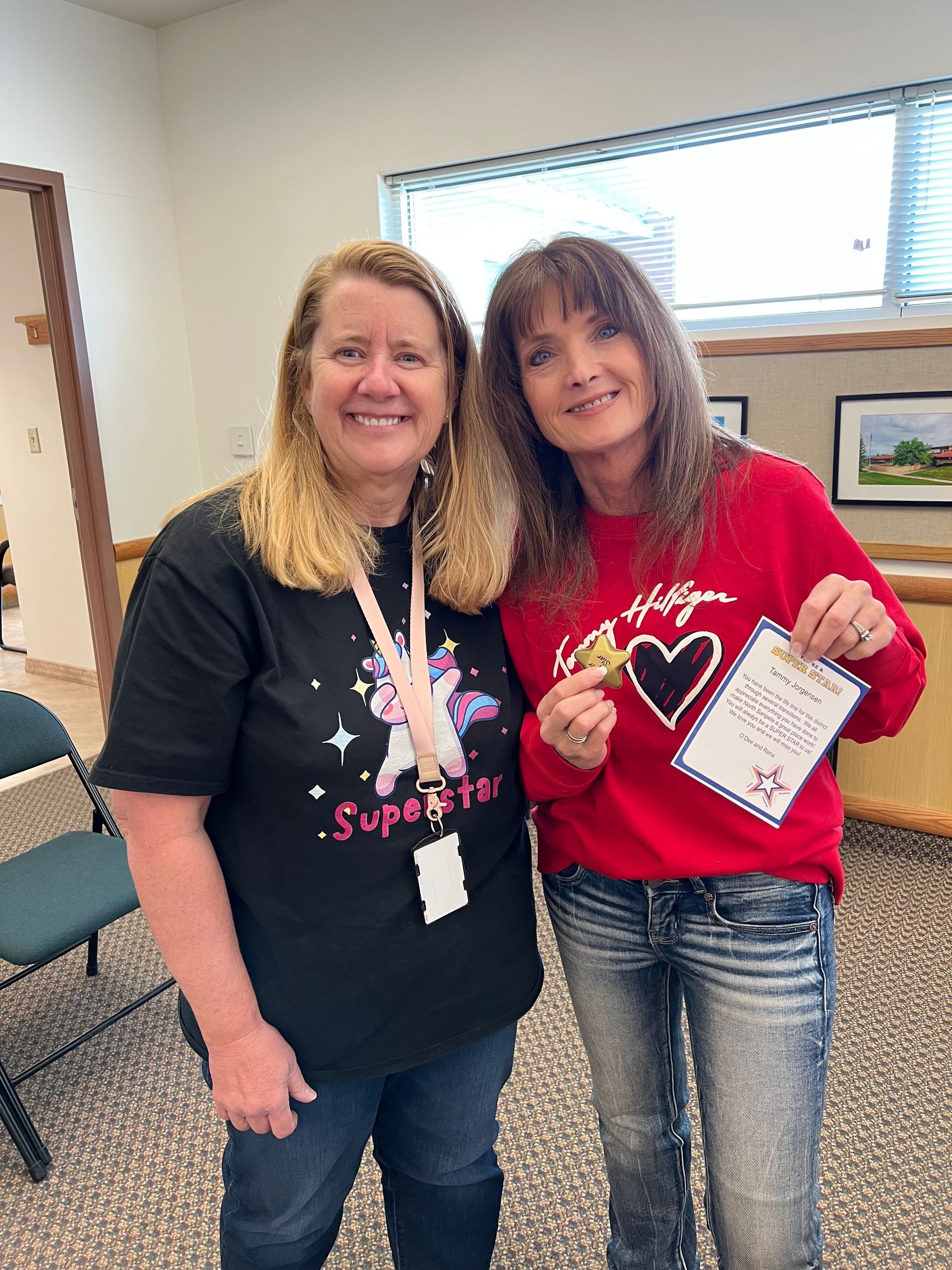 Two women smiling indoors, standing side by side and wearing heart-themed shirts with lanyards.