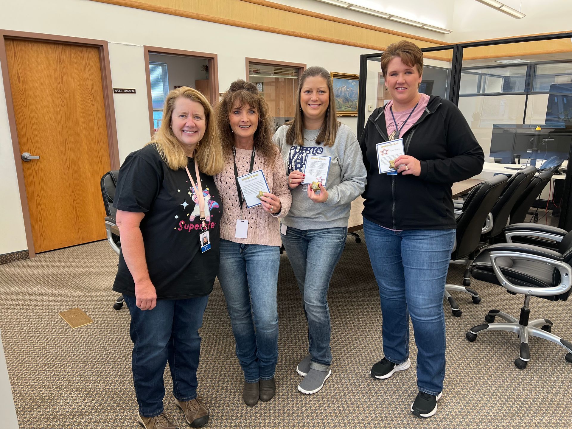 Four women in an office holding small cards, smiling. Jeans, casual tops. Background: office furniture, neutral colors.