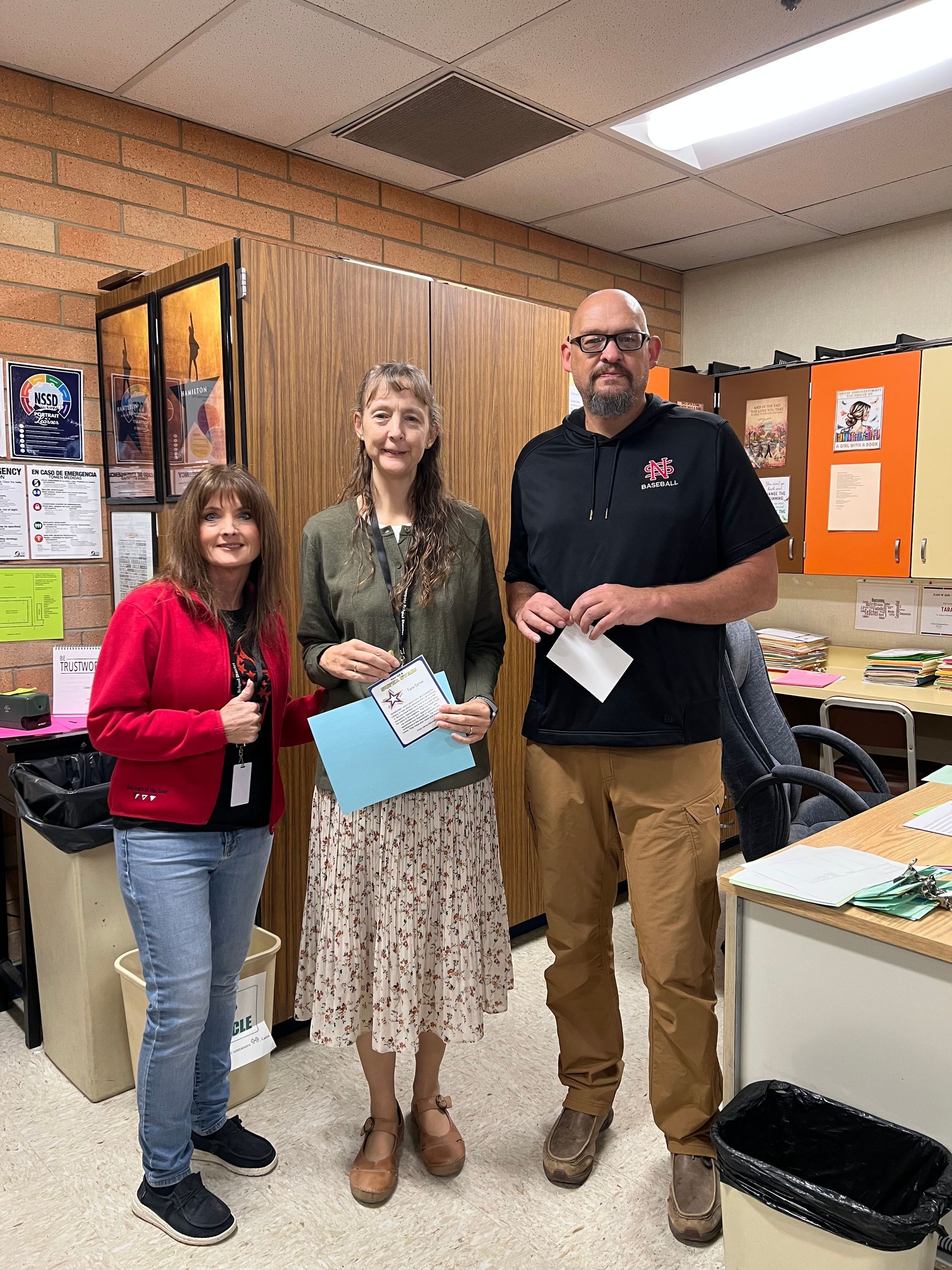 Three people standing in a classroom. Woman in red jacket, woman in floral skirt, man in black shirt.