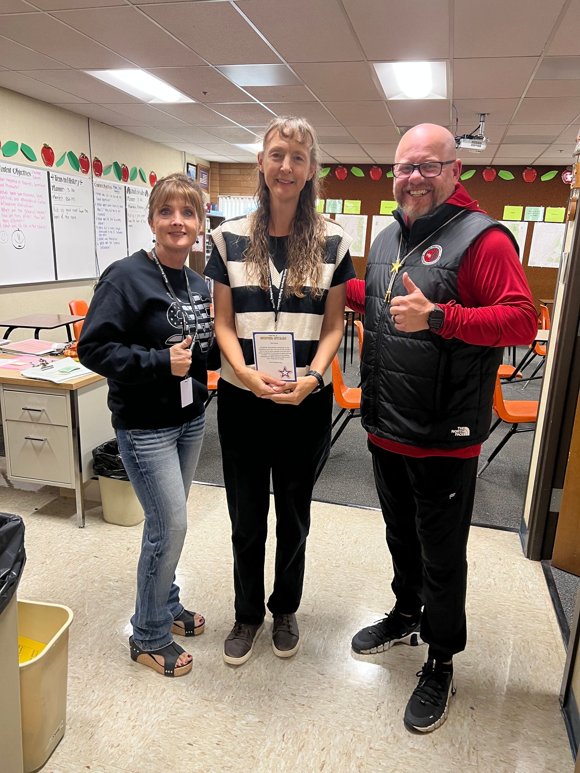 Three people in a classroom: two women and a man. Man gives a thumbs-up.