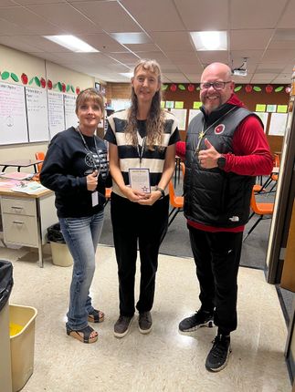 Three adults in a classroom. Woman in black shirt holds a pen, a woman in stripes, and a man in red vest gives a thumbs-up.