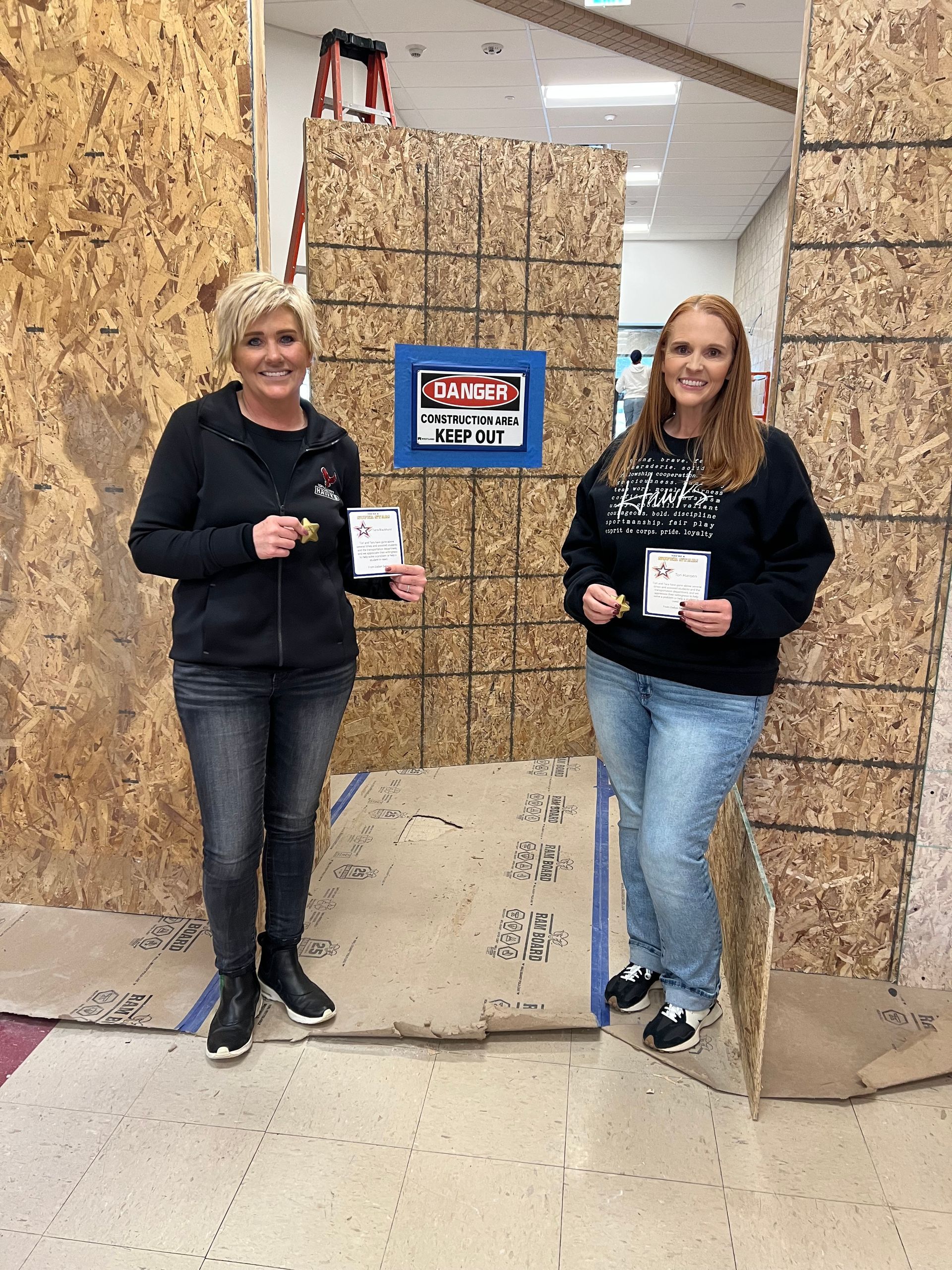 Two women holding keys stand in front of a plywood wall with a 