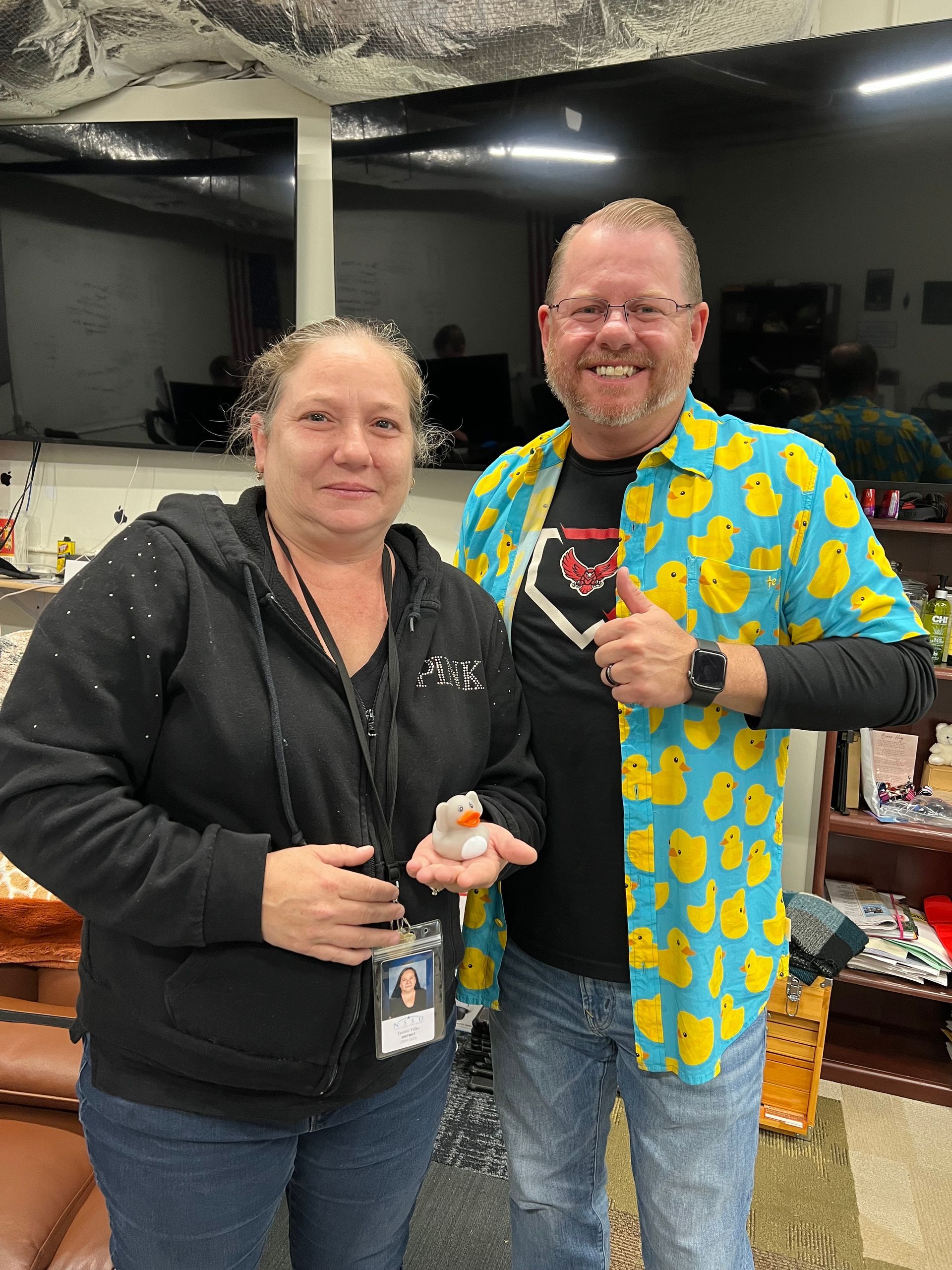 A man and a woman are posing for a picture in a living room . the man is wearing a duck shirt.