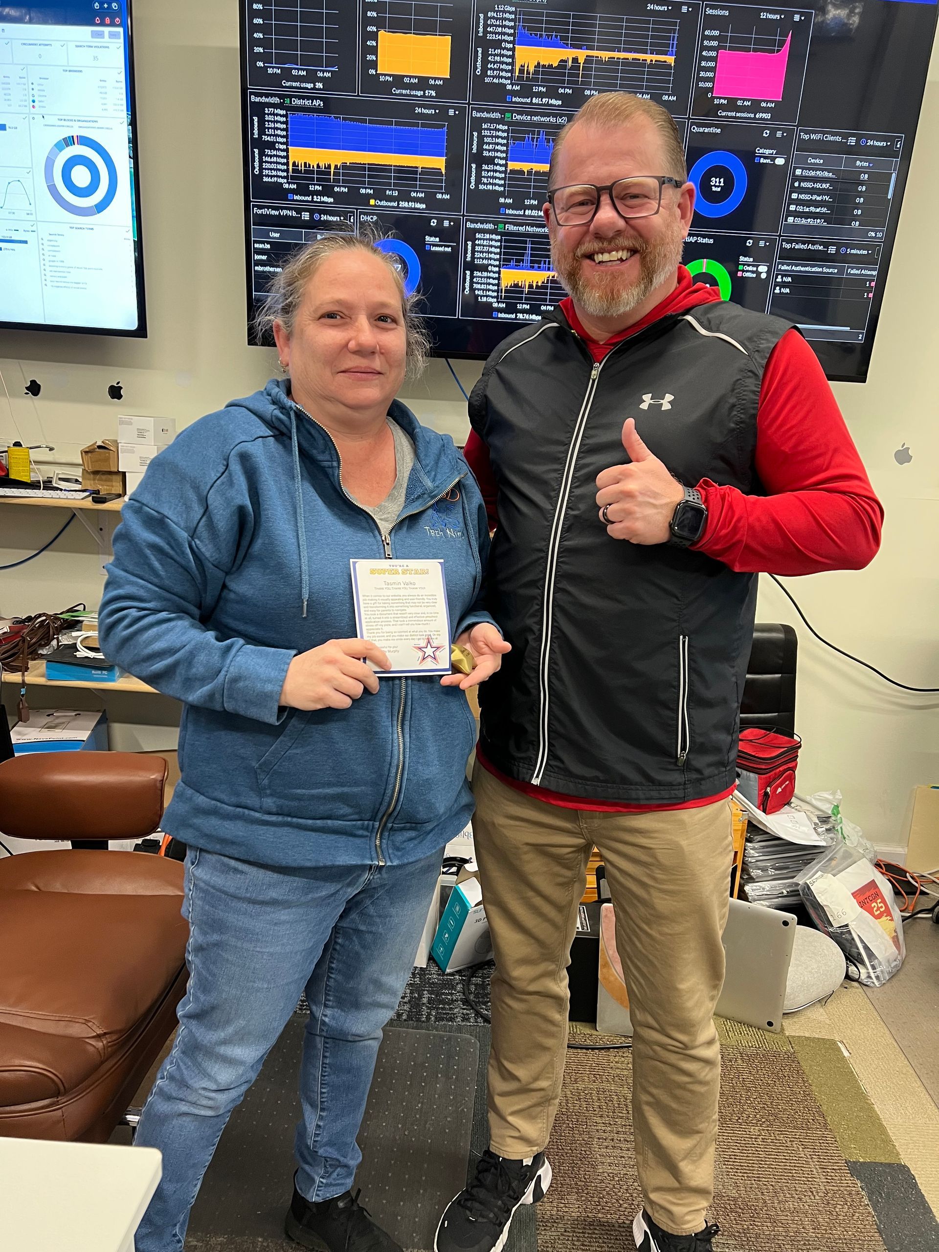 Two people stand in an office with computer screens, one holding a small note, the other giving a thumbs-up.