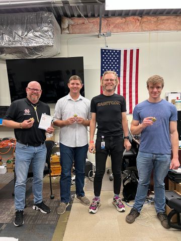 Four men hold snacks, posing in a workshop, American flag backdrop.