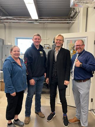 Four people standing indoors; two men, two women. They are smiling, and are in a mechanical-looking room.