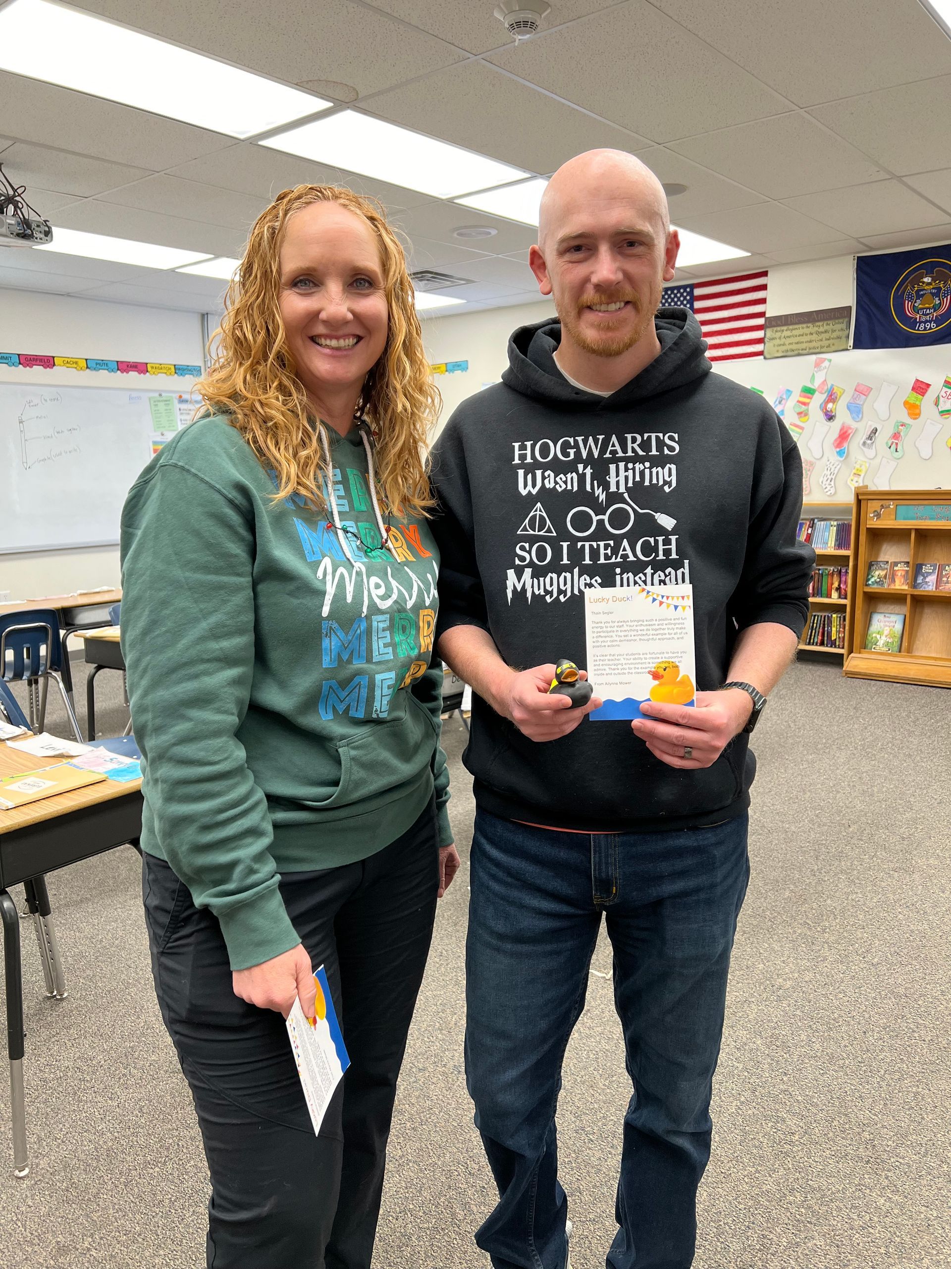 A man and a woman are standing next to each other in a classroom holding a book.