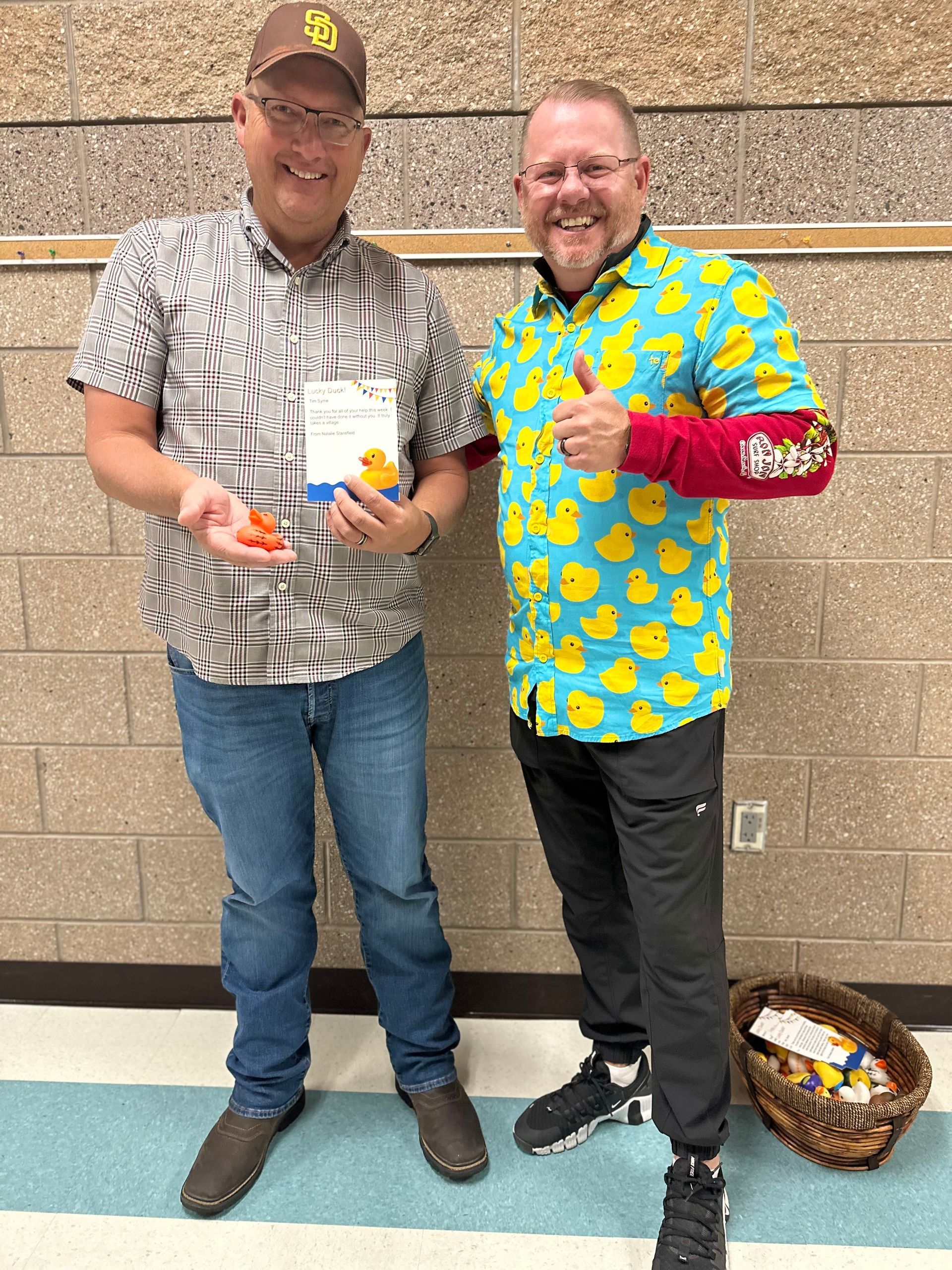 Two men are standing next to each other in front of a brick wall . one of the men is wearing a duck shirt.