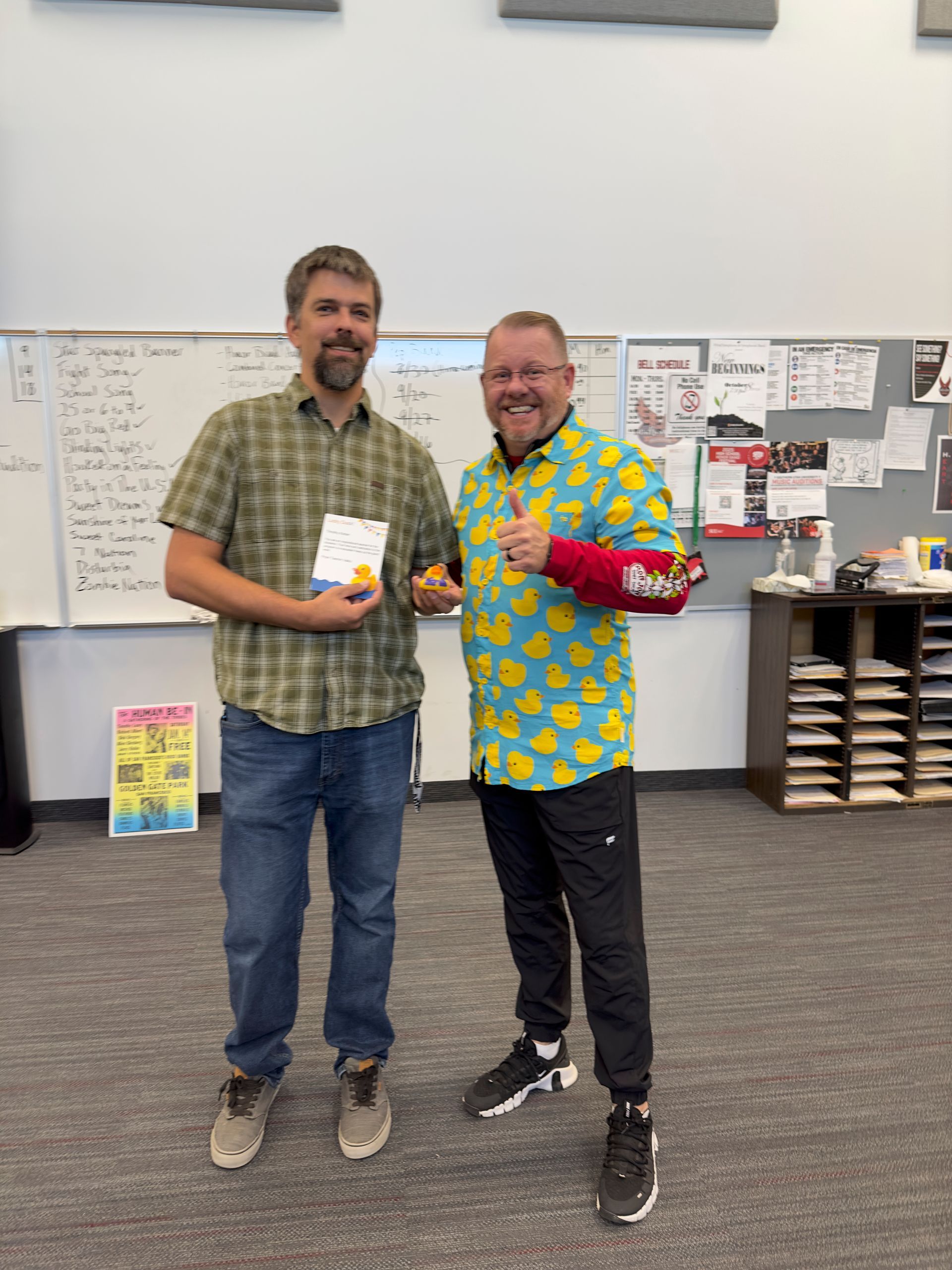 Two men are standing next to each other in a classroom holding a book.