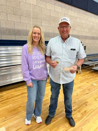 Two people standing in a gym, smiling and holding drinks near bleachers