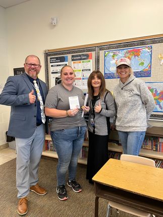 Four people stand in a classroom; a man gives a thumbs-up. Two women hold papers. A world map is in the background.
