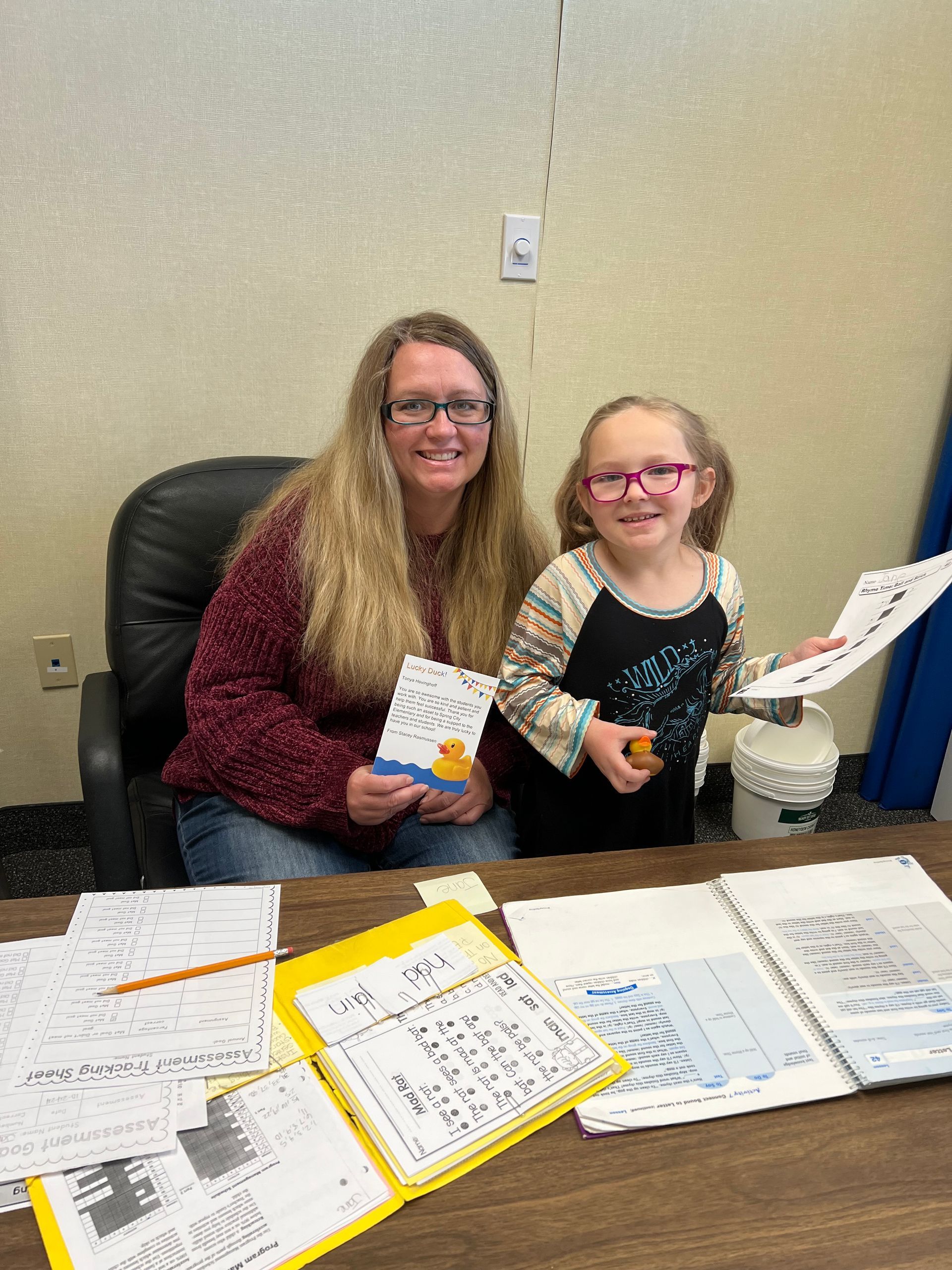 A woman and a little girl are sitting at a desk holding papers.