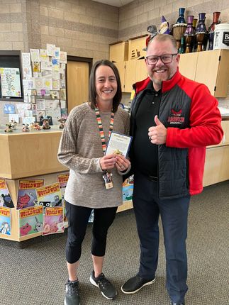 Two people smiling indoors, holding a certificate and giving a thumbs-up near gift displays.