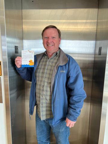 A man in a blue jacket is standing in an elevator holding a book.