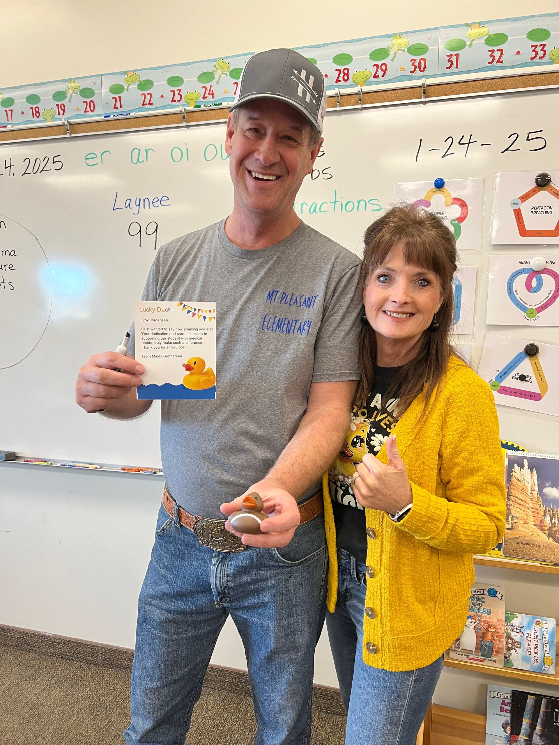 A man and a woman are standing next to each other in a classroom holding a book.