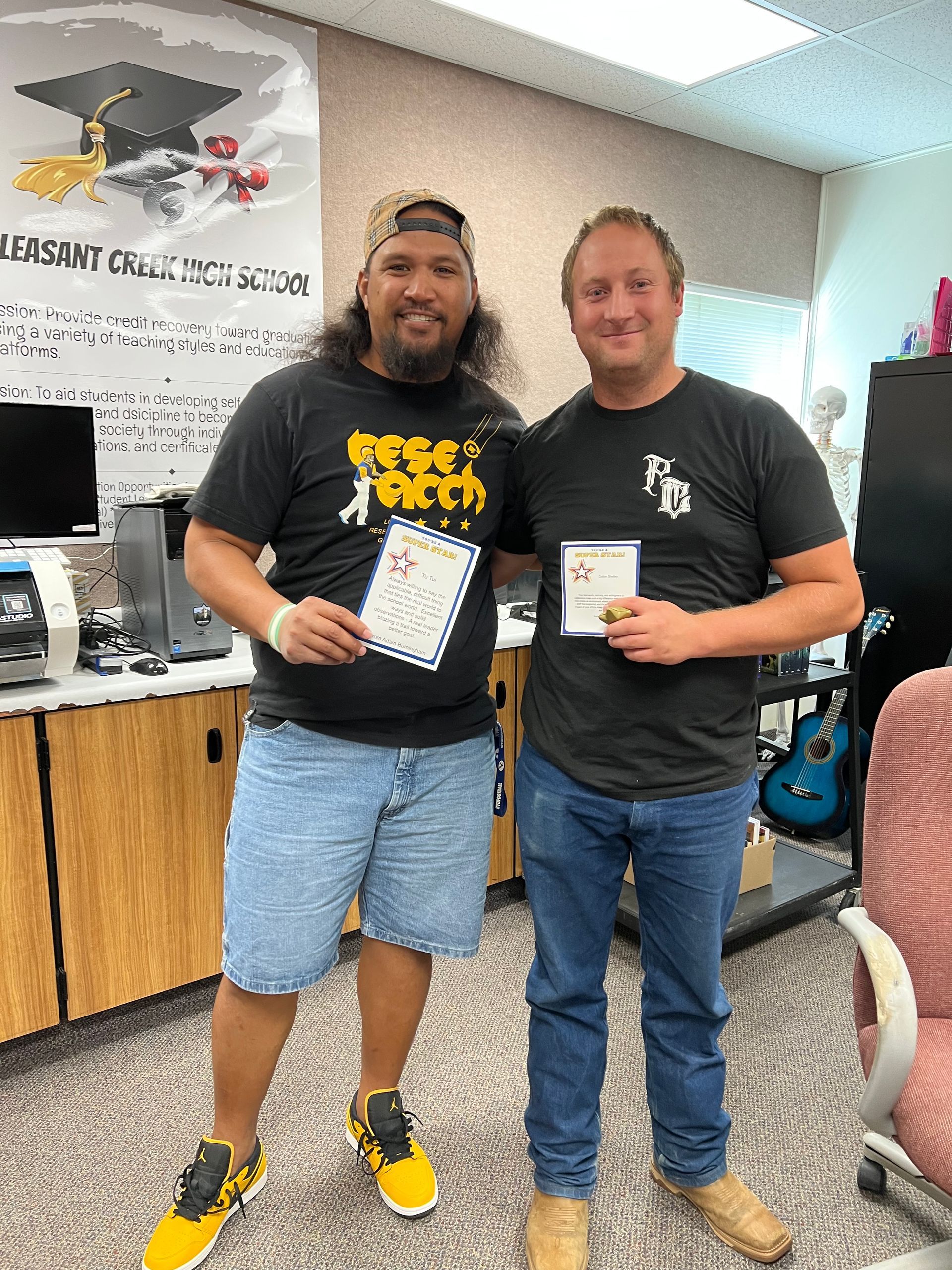 Two men holding certificates smile in an office setting. One in shorts, the other in jeans.