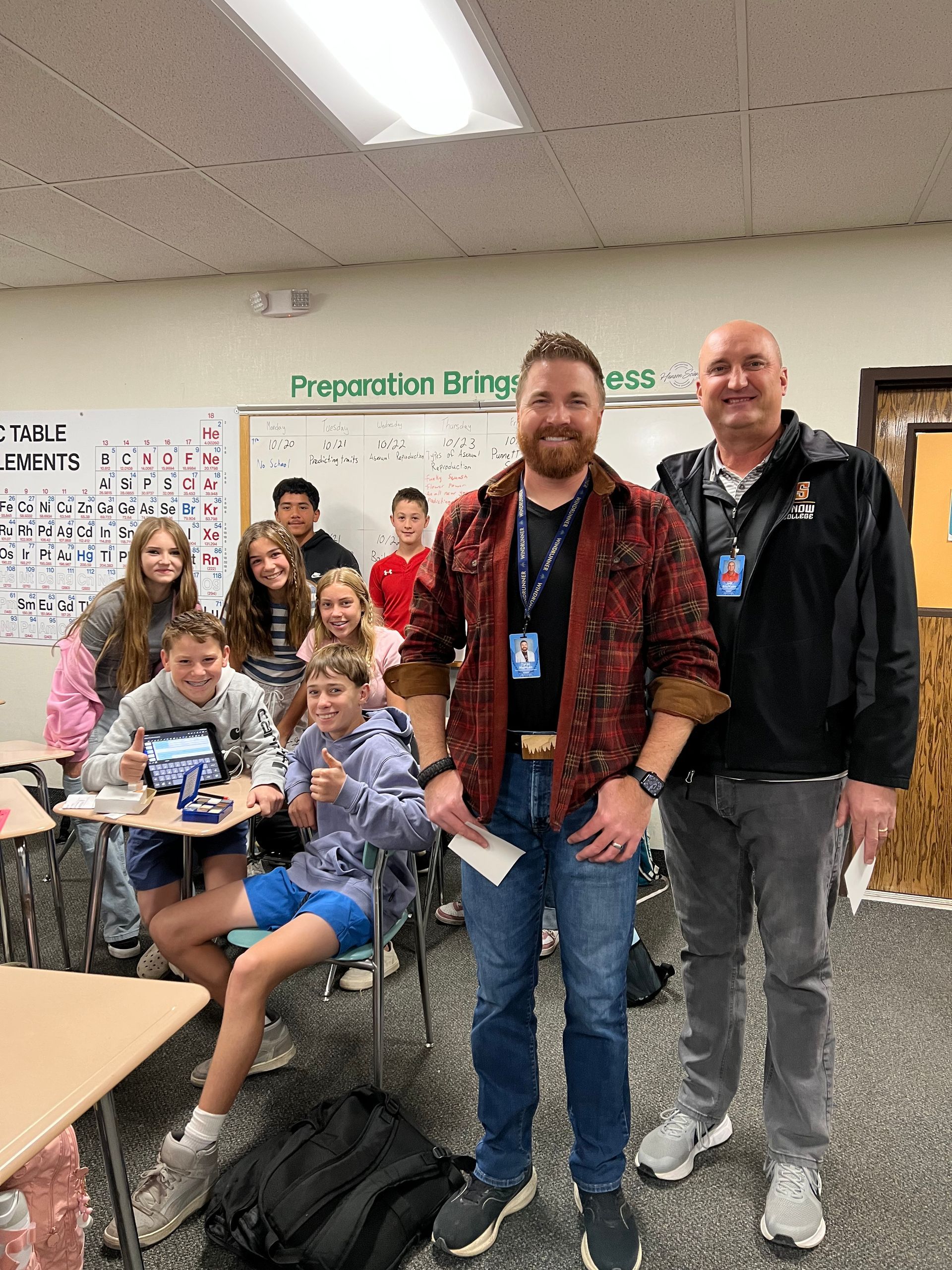 Teacher and man stand in a classroom with smiling students. Whiteboard in background.