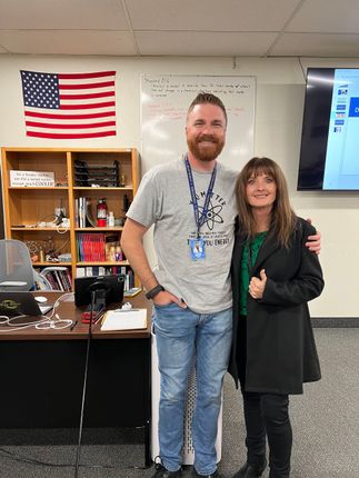 Two people standing close together in an office. Man in grey t-shirt and jeans, woman in black coat. American flag on wall.
