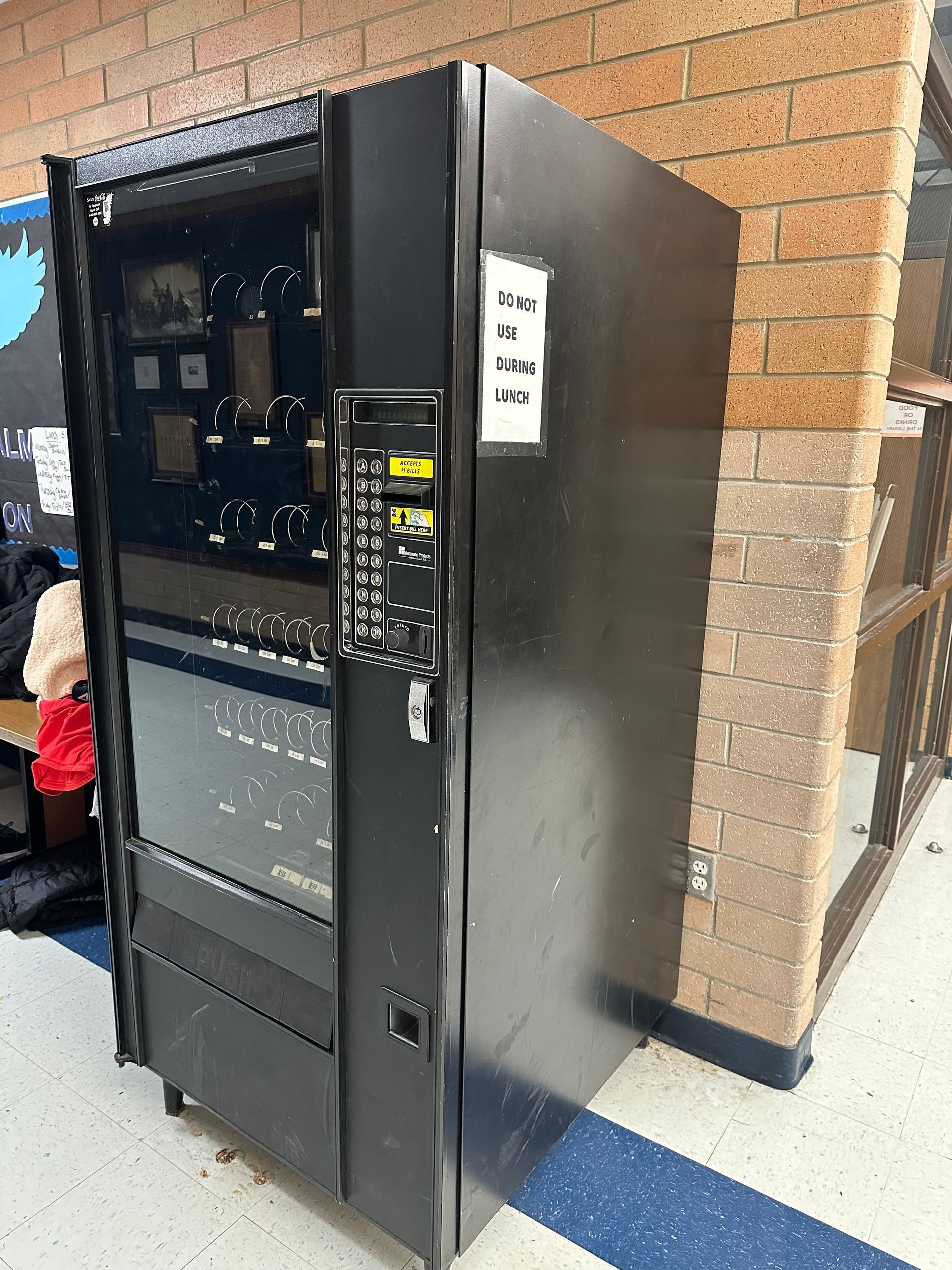 A black vending machine is sitting in a hallway next to a brick wall.