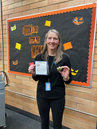 Woman holding sign and small object, standing in front of bulletin board with Halloween decorations.