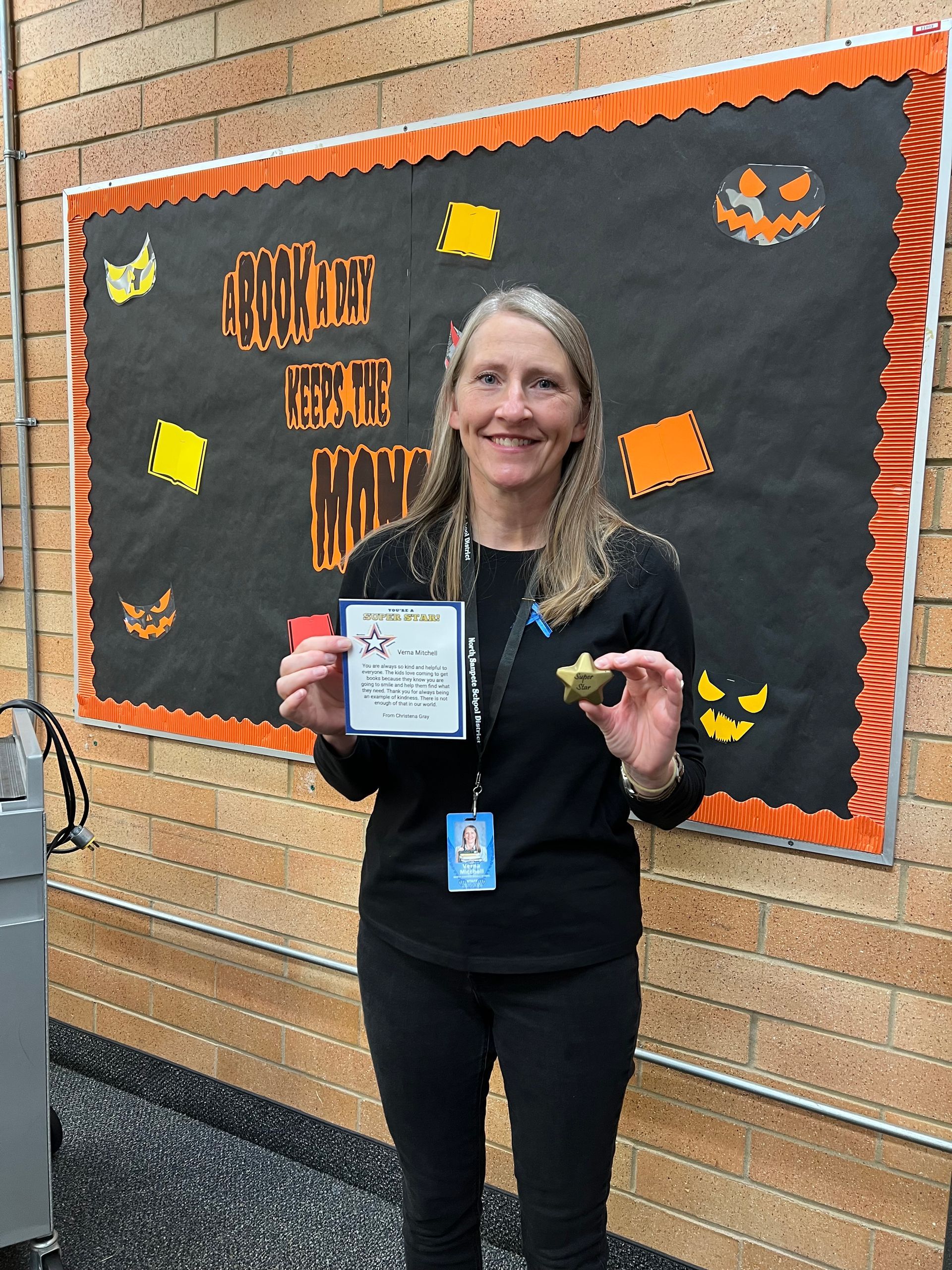 Woman holding a sign and a small item, standing in front of a Halloween bulletin board.