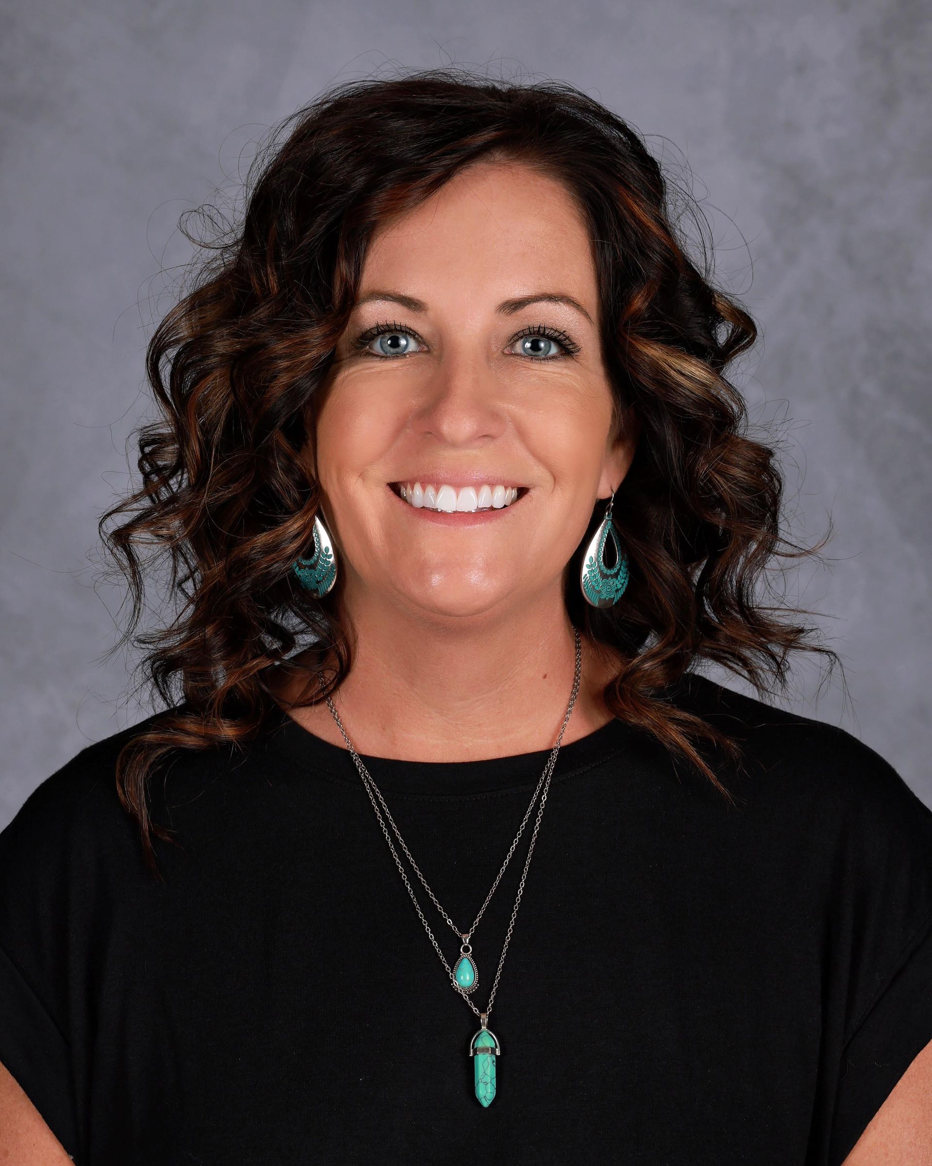 A smiling woman with dark curly hair wearing a black top and turquoise pendant necklace against a gray background.