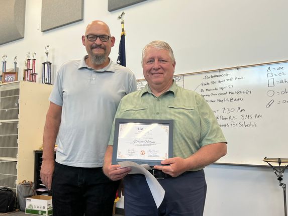 Two men stand in a classroom, one holding a framed certificate and smiling beside a whiteboard.