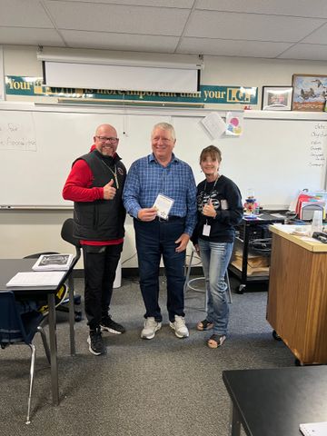 Three people pose in a classroom; two men and a woman. They are smiling and holding objects.
