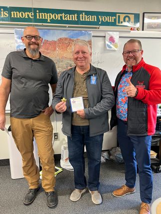 Three men smiling indoors, posing with a certificate under a sign that says “is more important than your IQ.”