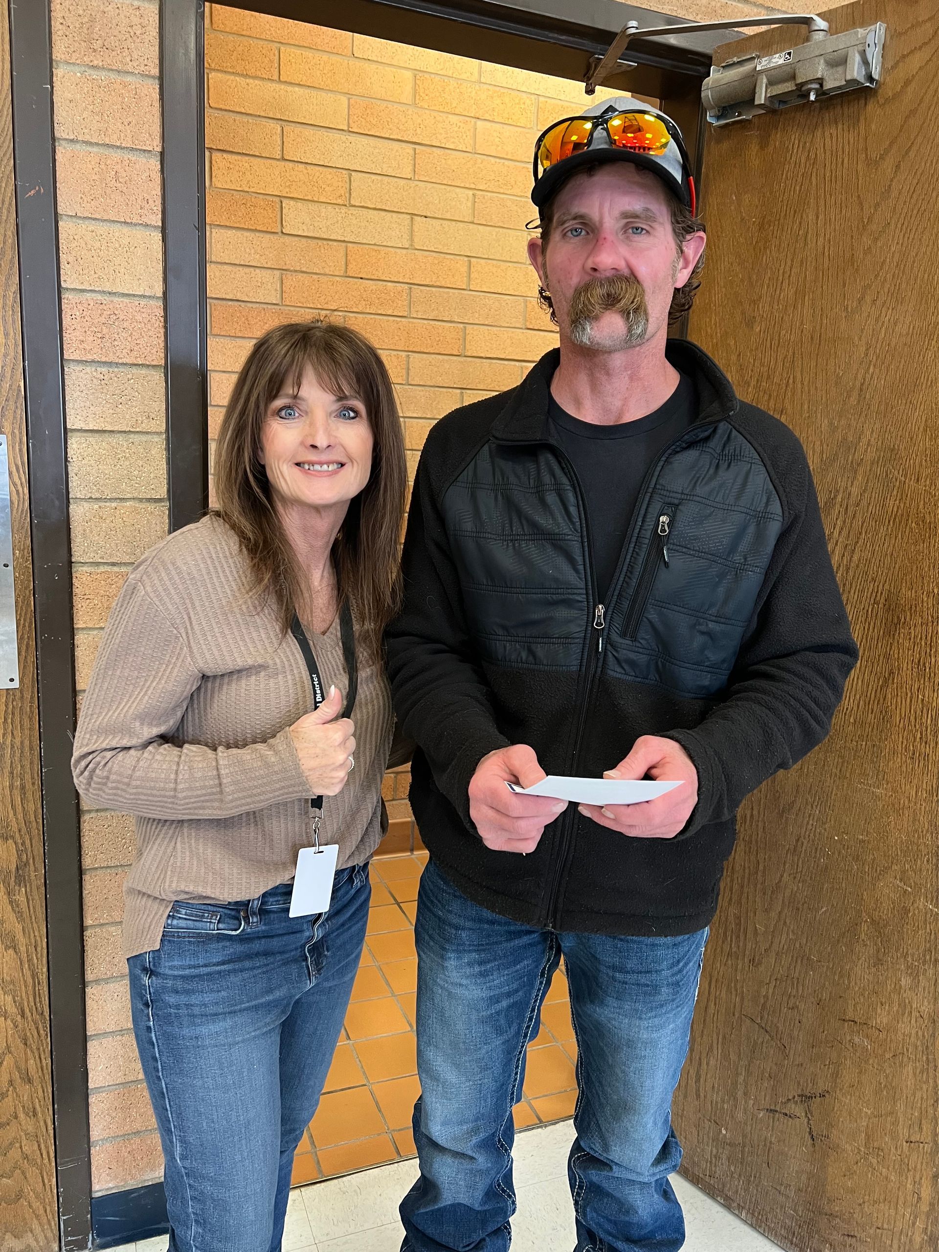A person in a beige sweater and a person in a black jacket and cap stand together in front of a brick wall.