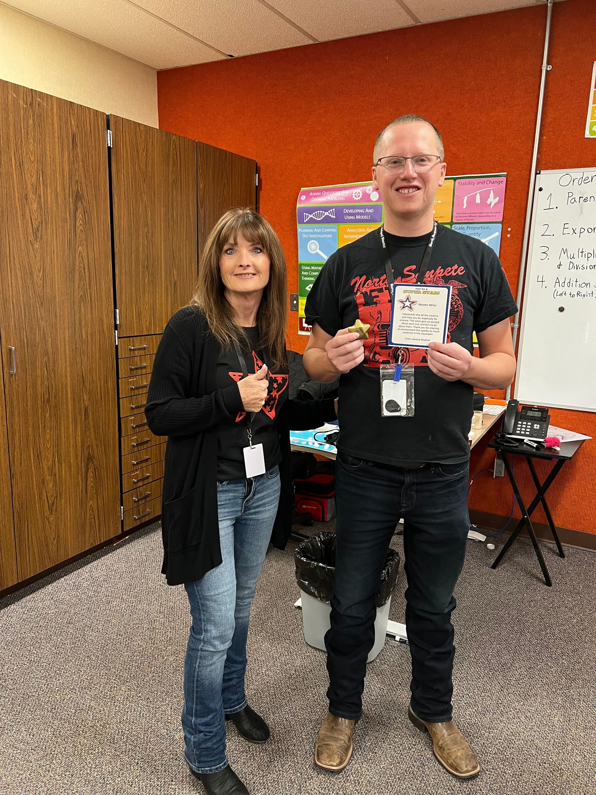 Two people standing in a classroom; a woman wearing a black cardigan and jeans, a man holding something, both wearing black shirts.
