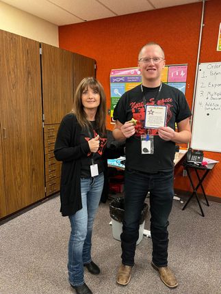 Two people stand in a room, the man holding a small object. They both wear black shirts. Orange and brown interior.