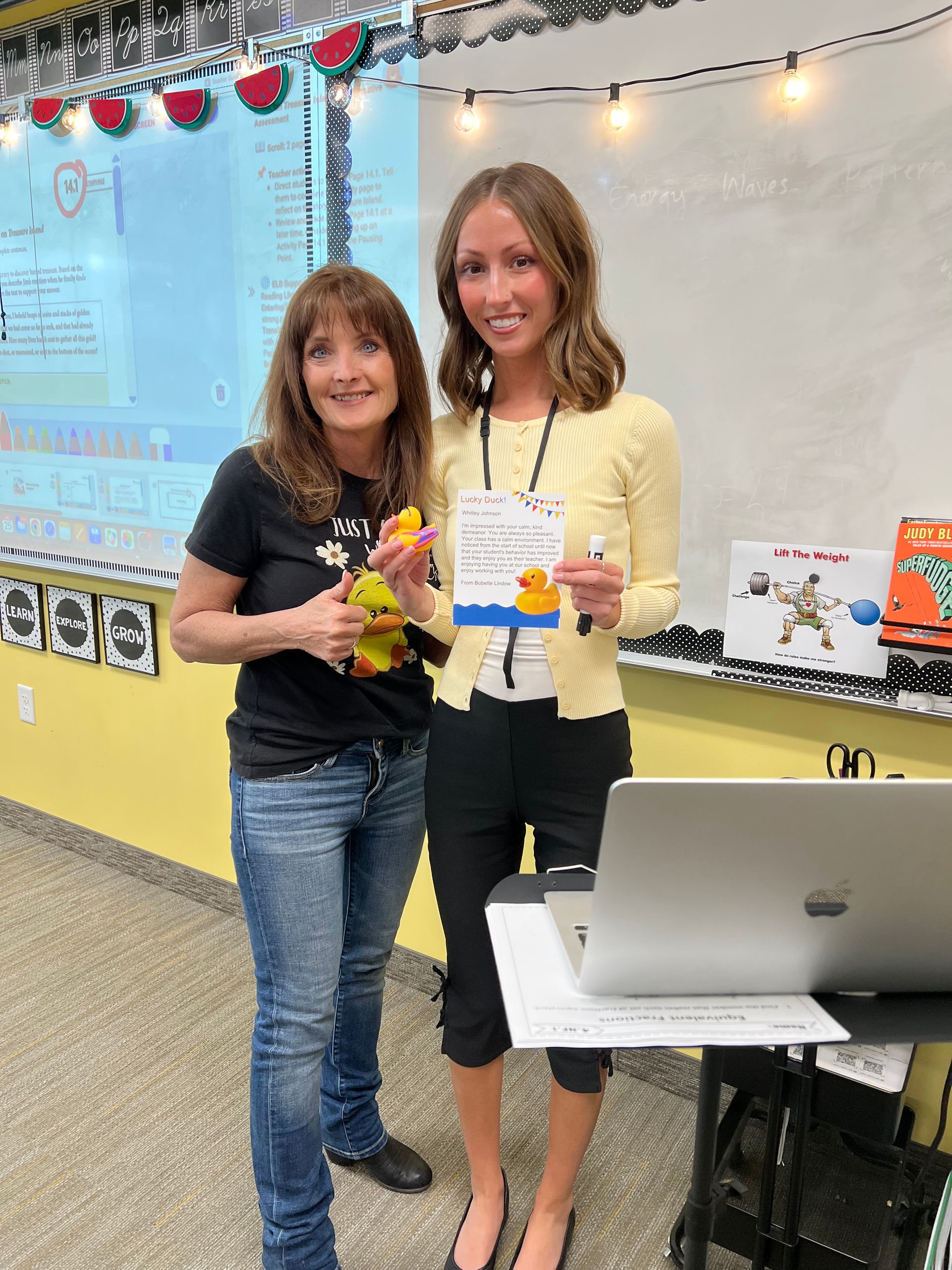 Two women are standing next to each other in front of a laptop in a classroom.
