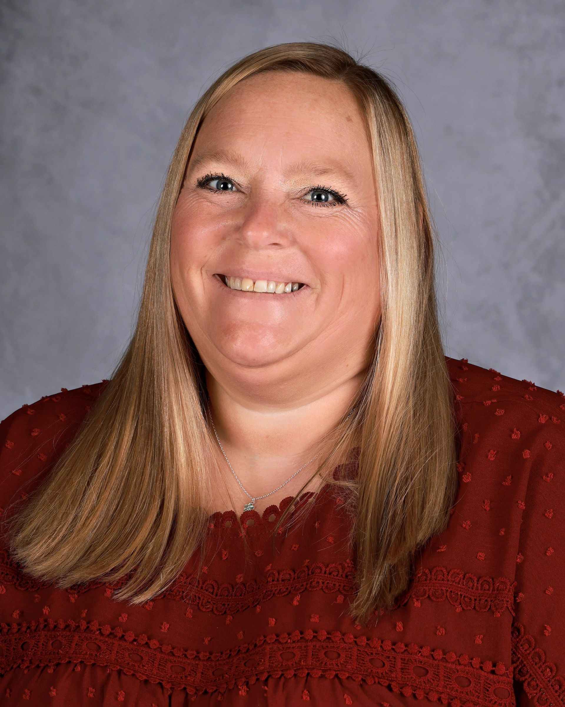 Smiling woman with long blonde hair wearing a rust-colored blouse against a grey backdrop.