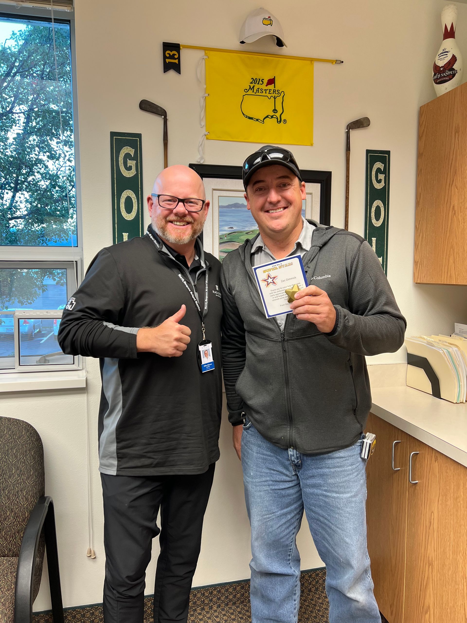Two men standing in an office, one giving a thumbs up. Golf-themed decorations on the wall.