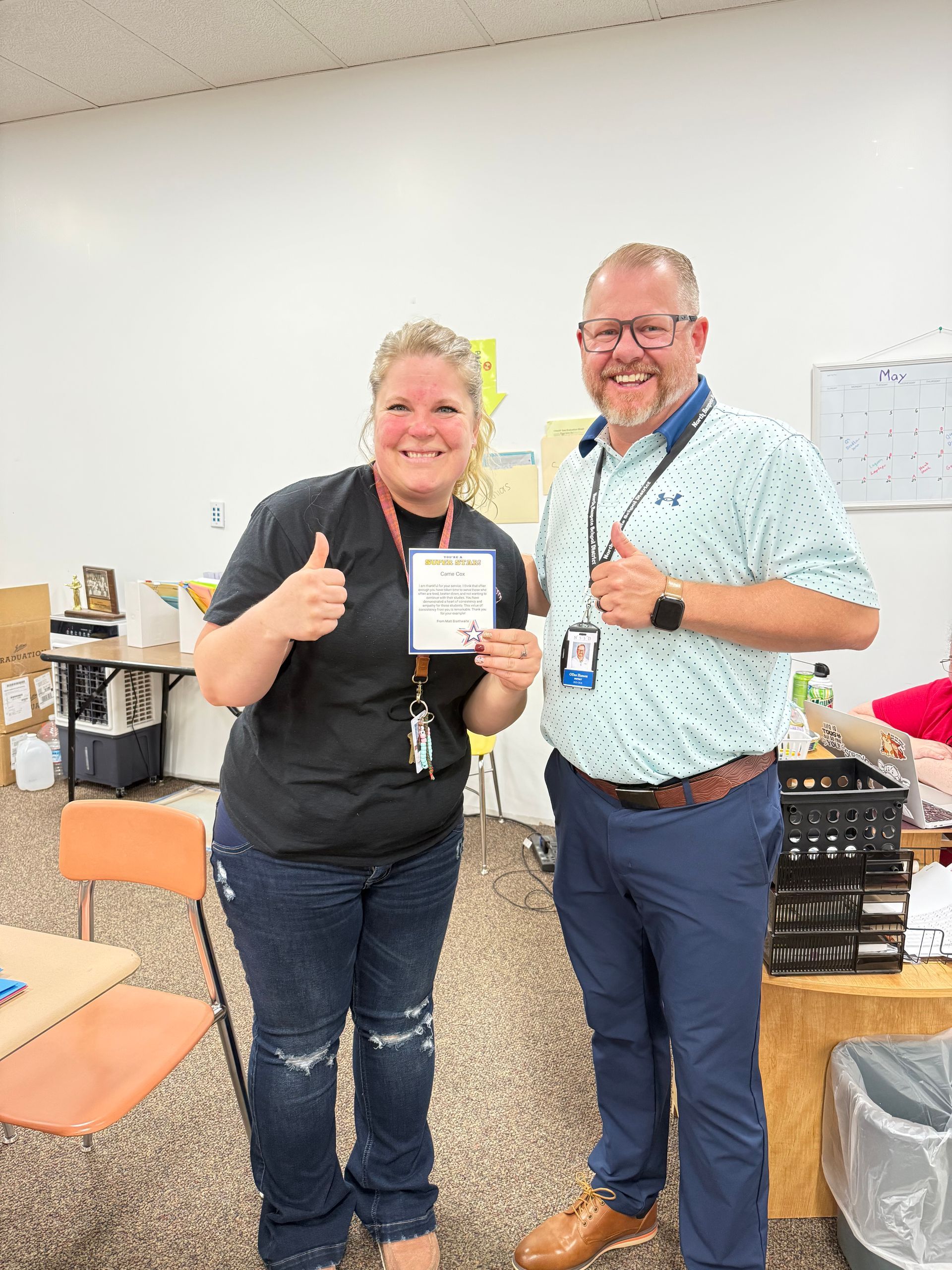 Two smiling coworkers posing indoors, holding documents and giving thumbs up near office equipment.