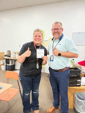 Two smiling people in a classroom, holding a certificate and giving thumbs up.